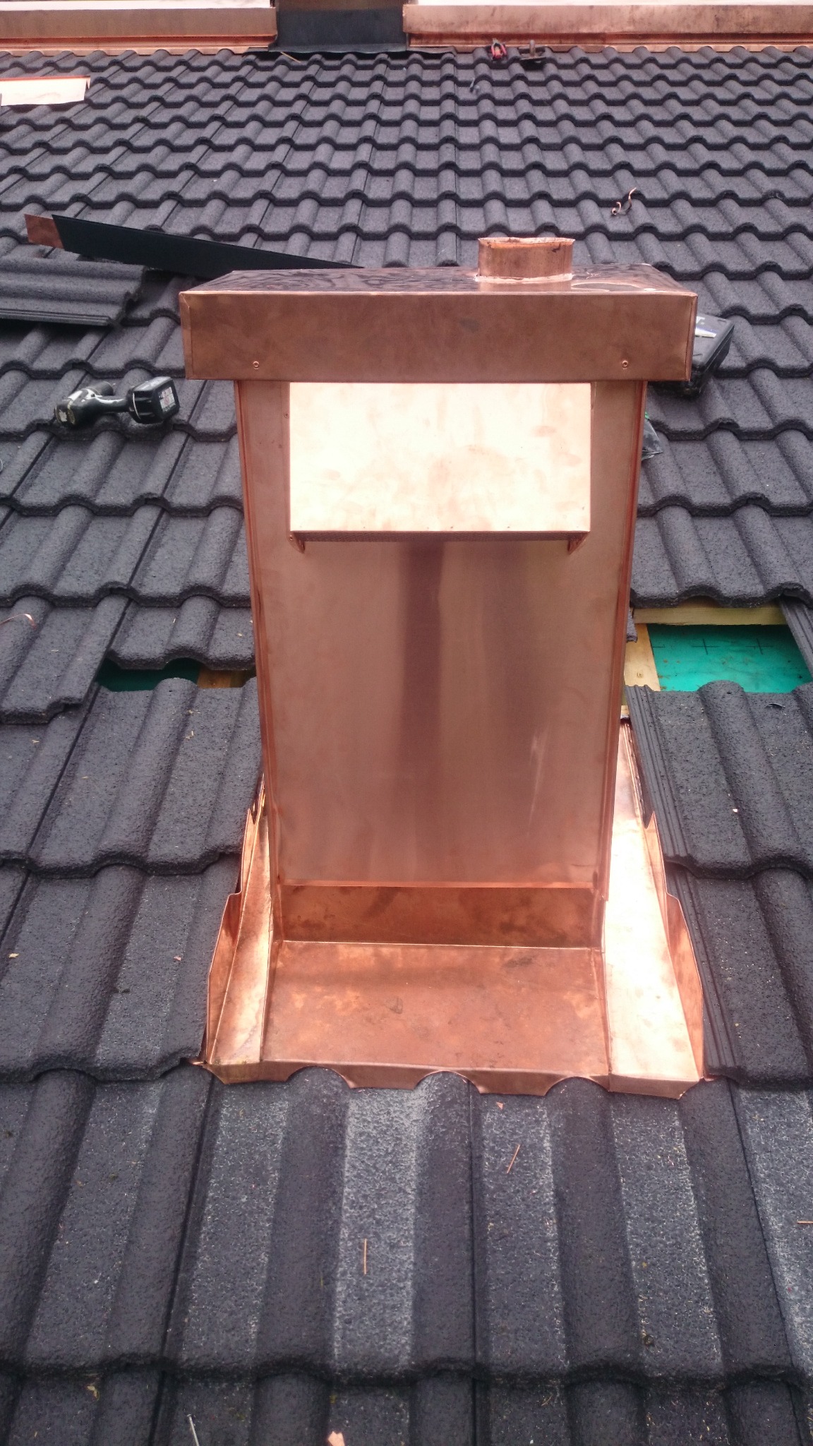 Miedziana obróbka komina na dachu z ciemnej dachówki bitumicznej, widoczne narzędzia i fragmenty niezakończonej pracy.