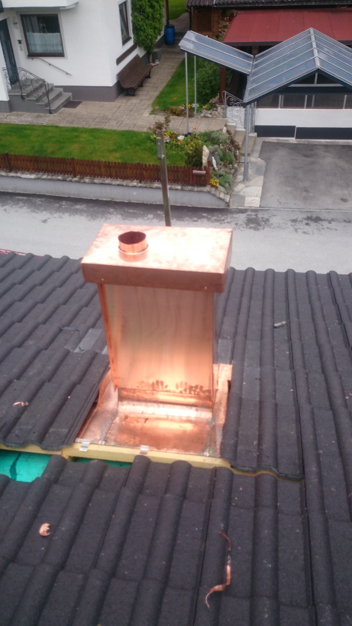 Miedziana obróbka komina na dachu z ciemnej dachówki ceramicznej, widok z góry na dom z ogrodem.