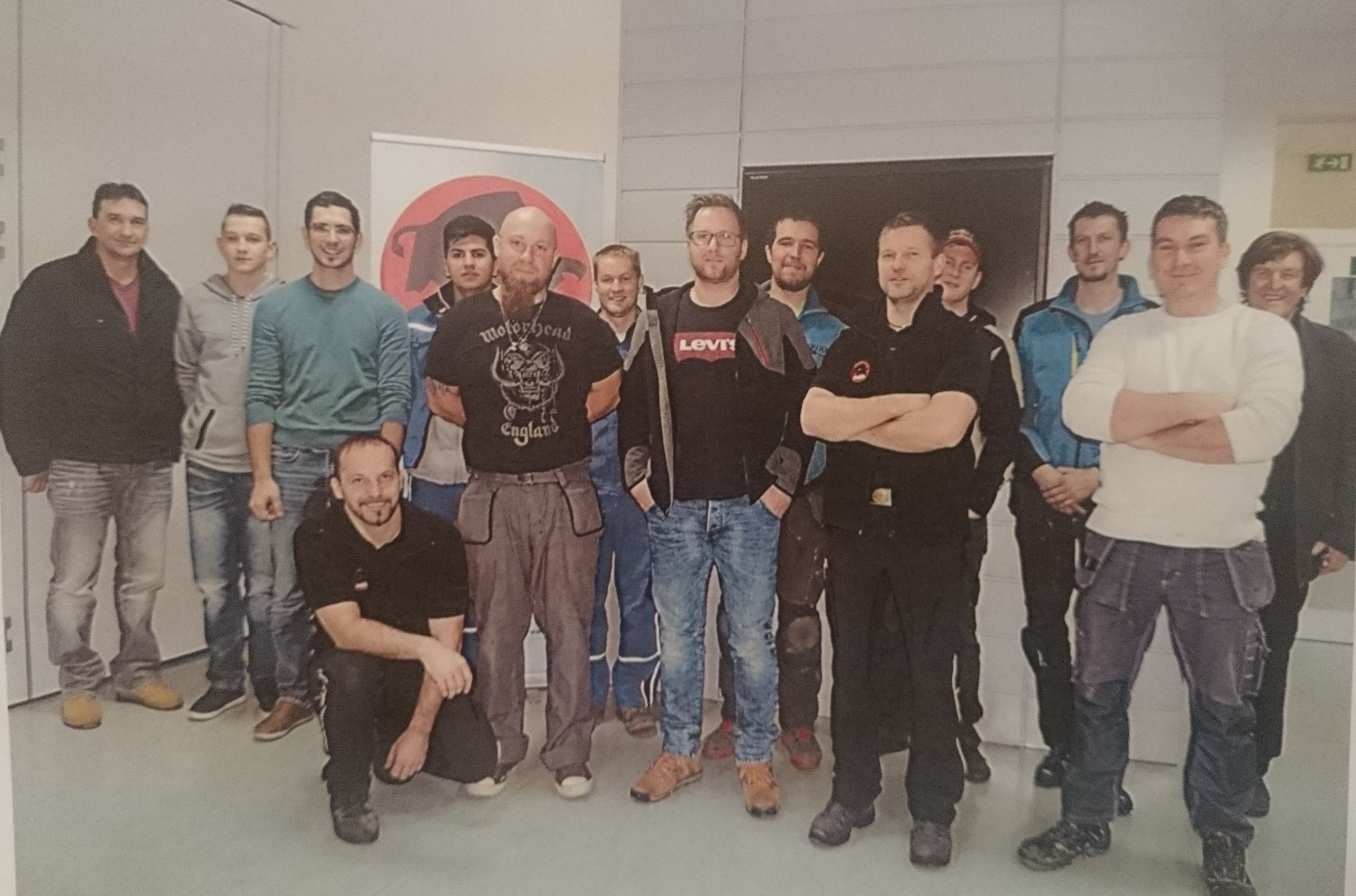 Grupa dekarzy uczestnicząca w szkoleniu, w tle widoczny baner z logo firmy, część z nich ubrana w odzież roboczą.