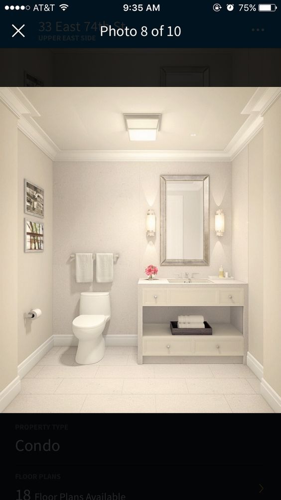 Jasna łazienka w stylu klasycznym z toaletą, umywalką z szafką, lustrem w srebrnej ramie, dwoma kinkietami i dekoracjami kwiatowymi.