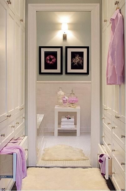 Wnętrze garderoby z białymi szafami, puchatym dywanem i dekoracjami w odcieniach różu, oświetlone kinkietem nad dwoma obrazami w czarnych ramkach.
