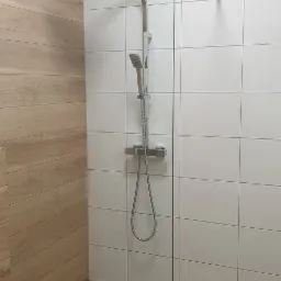 Nowoczesna kabina prysznicowa walk-in z odpływem liniowym, wykończona płytkami imitującymi drewno i białymi płytkami kwadratowymi.