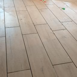 Bud Man - Ułożone płytki podłogowe imitujące drewno z widocznymi krzyżykami dystansowymi, w tle narzędzia i materiały budowlane.