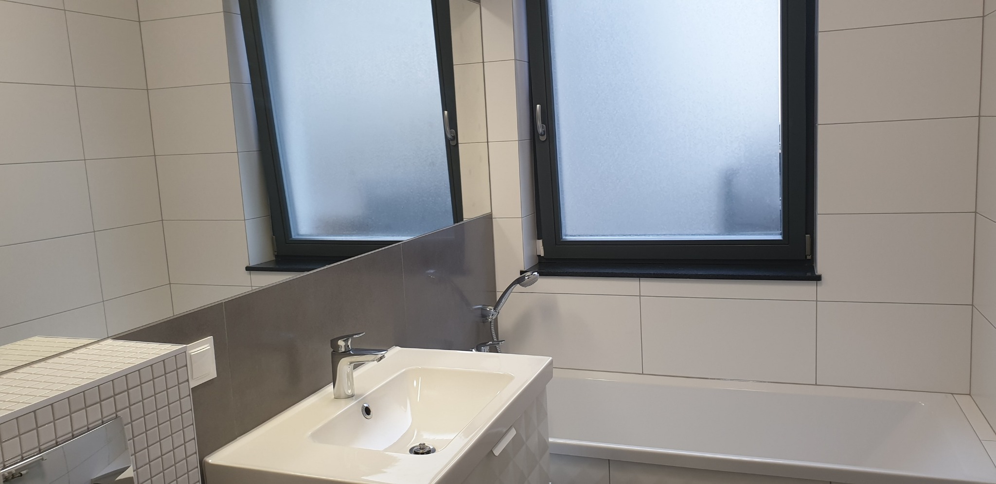 Nowoczesna łazienka z białymi płytkami, umywalką z szafką o geometrycznym wzorze, baterią chromowaną i dwoma oknami z matowymi szybami.