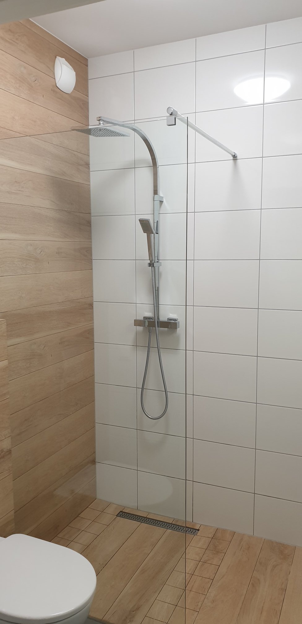 Nowoczesna kabina prysznicowa walk-in z odpływem liniowym, wykończona płytkami imitującymi drewno i białymi płytkami kwadratowymi.