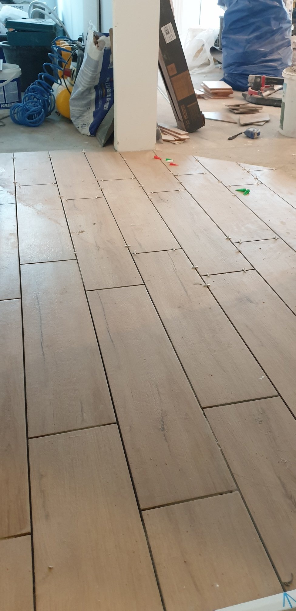 Ułożone płytki podłogowe imitujące drewno z widocznymi krzyżykami dystansowymi, w tle narzędzia i materiały budowlane.