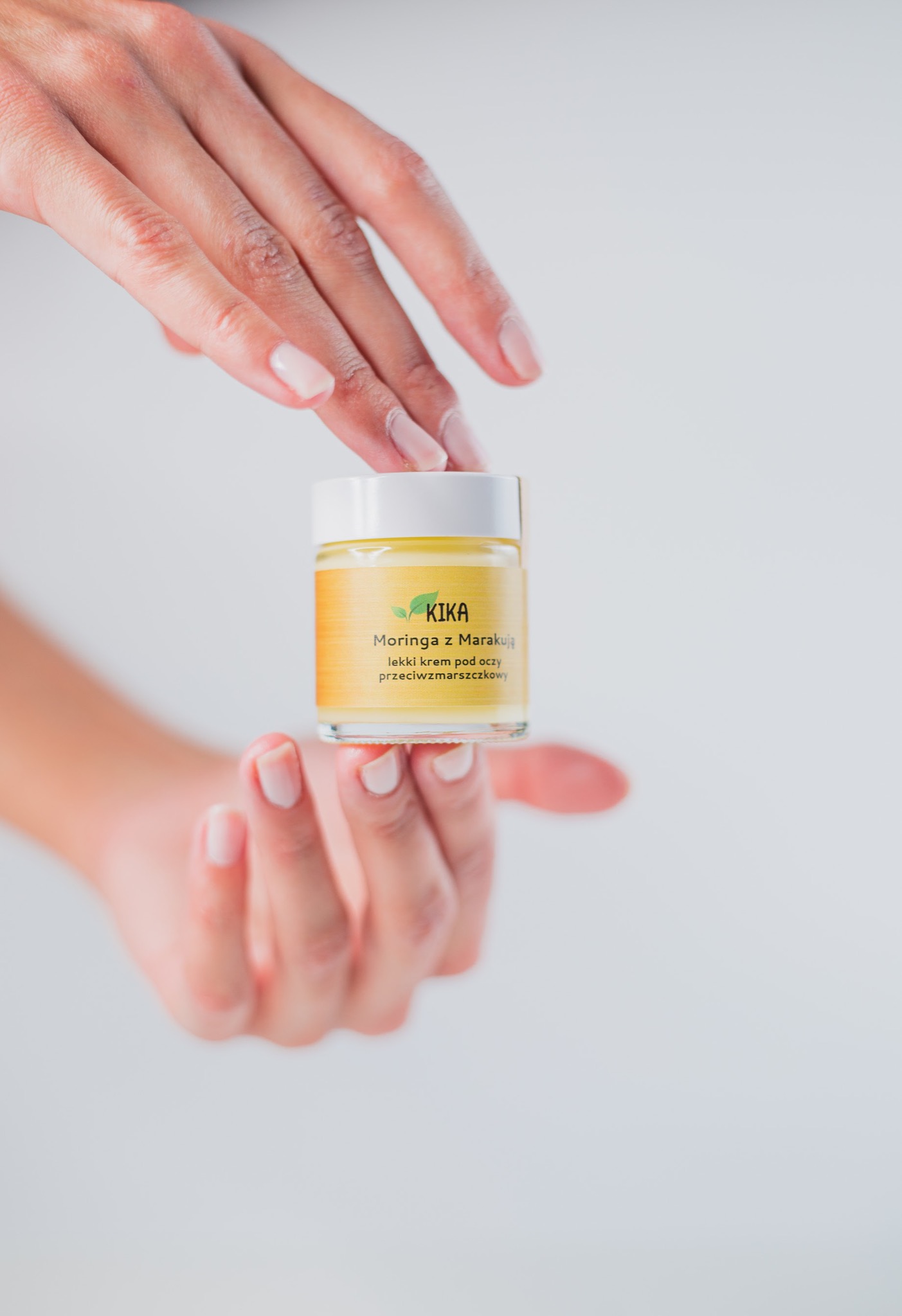 Dwie dłonie trzymają słoiczek kremu KIKA Moringa z Marakują, lekki krem pod oczy przeciwzmarszczkowy, na białym tle.