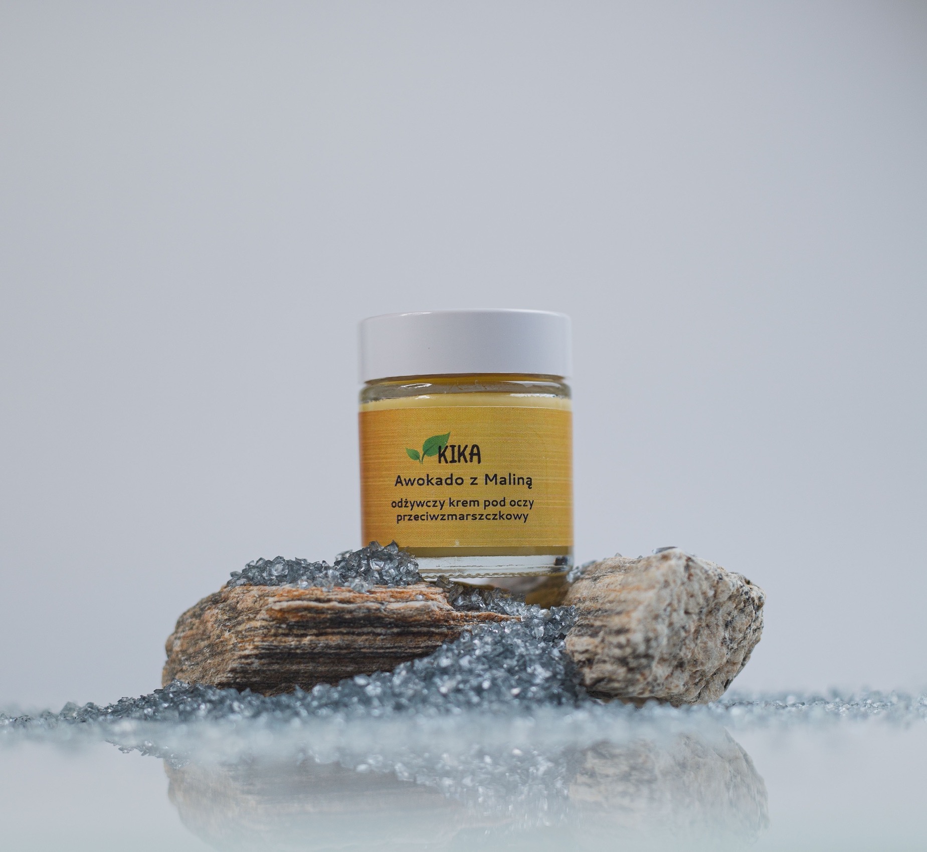 Słoik kremu pod oczy 'Awokado z Maliną' marki KIKA, umieszczony na kamieniach i szarych kryształkach na błyszczącym tle.