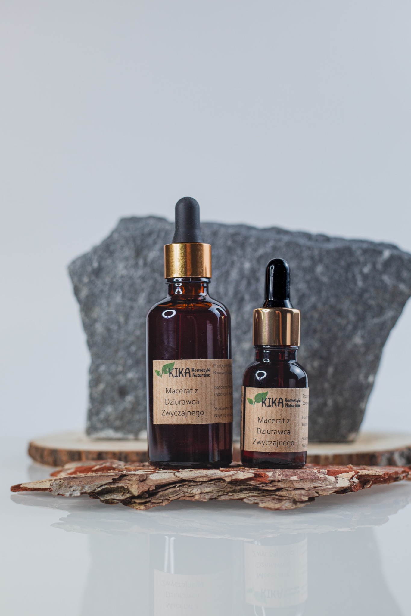 Dwie butelki z brązowego szkła z pipetami, zawierające macerat z dziurawca, stoją na kawałku kory drzewnej, z szarym kamieniem w tle.