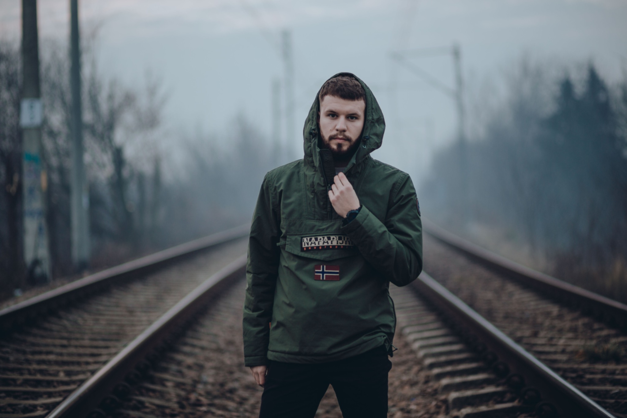 Mężczyzna w zielonej kurtce z kapturem stoi na torach kolejowych w mglisty dzień, patrząc w obiektyw.