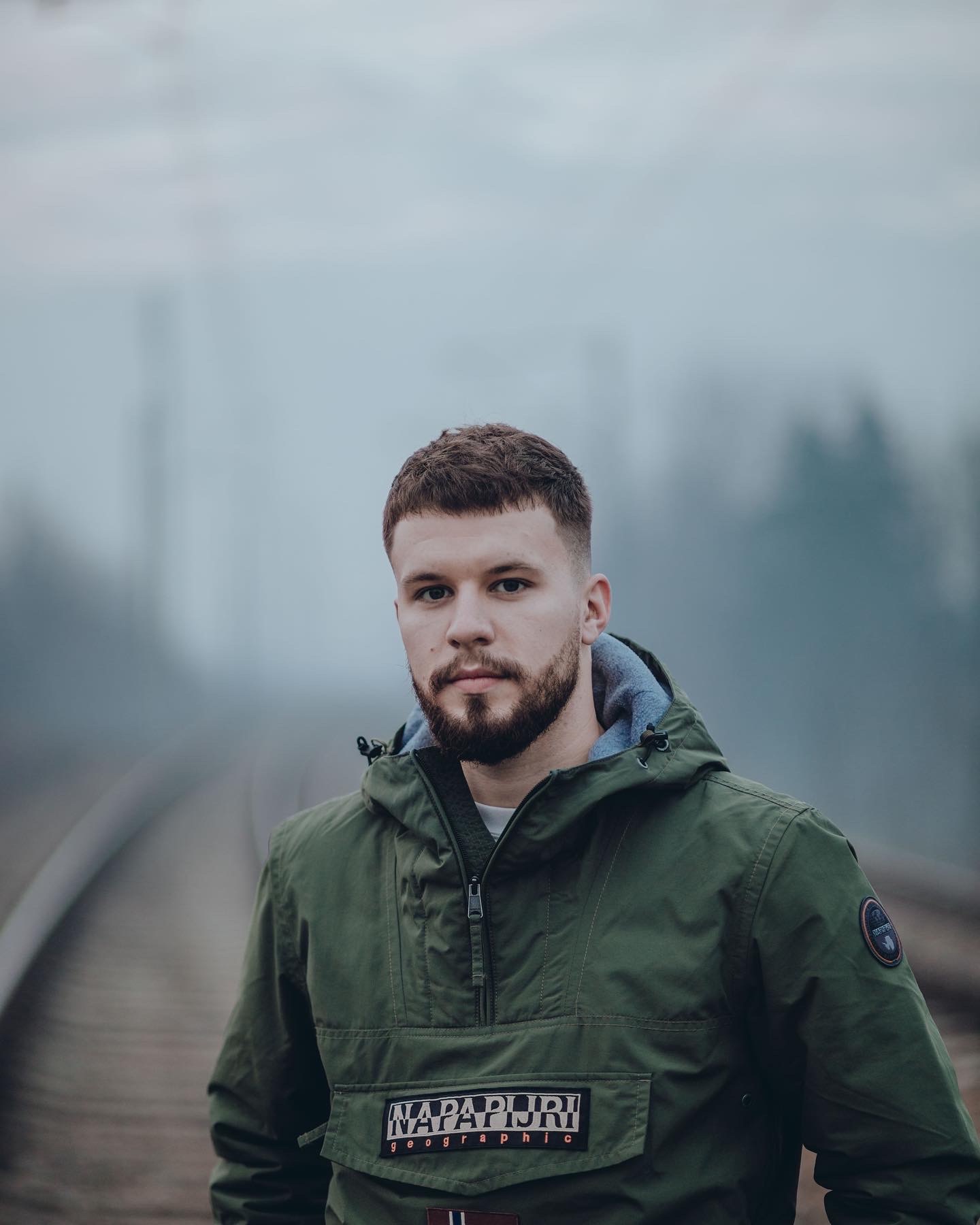 Młody mężczyzna z brodą w zielonej kurtce marki Napapijri, stojący na torach kolejowych we mgle.