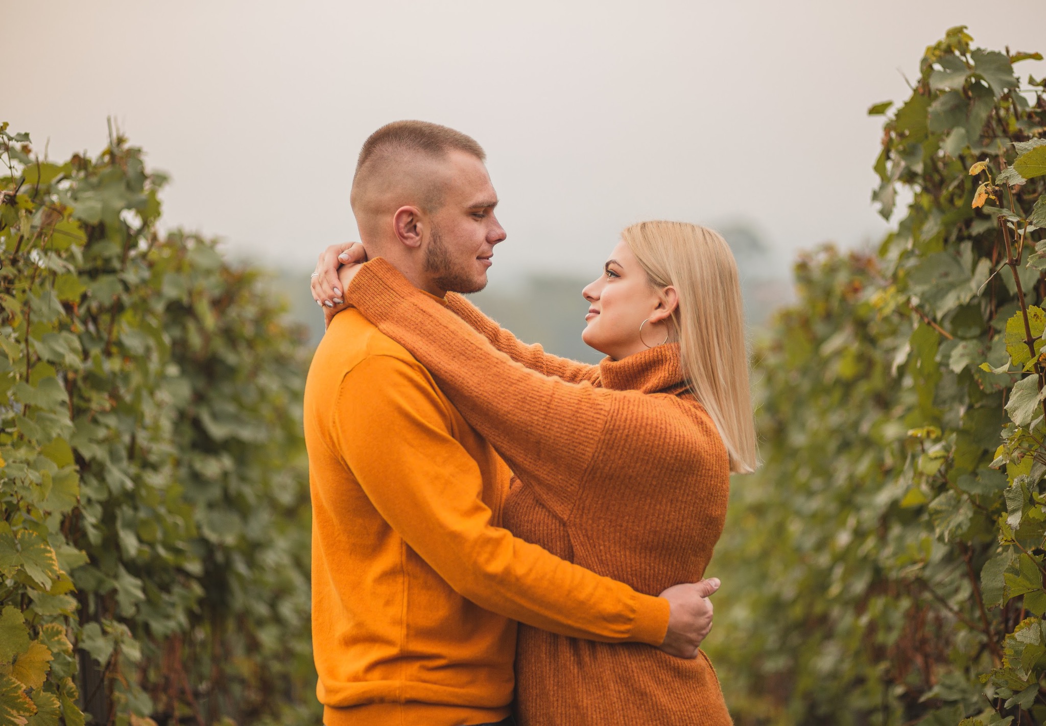 Para zakochanych obejmuje się w winnicy, oboje ubrani w pomarańczowe swetry, ona z kolczykami kołami i pierścionkiem zaręczynowym.