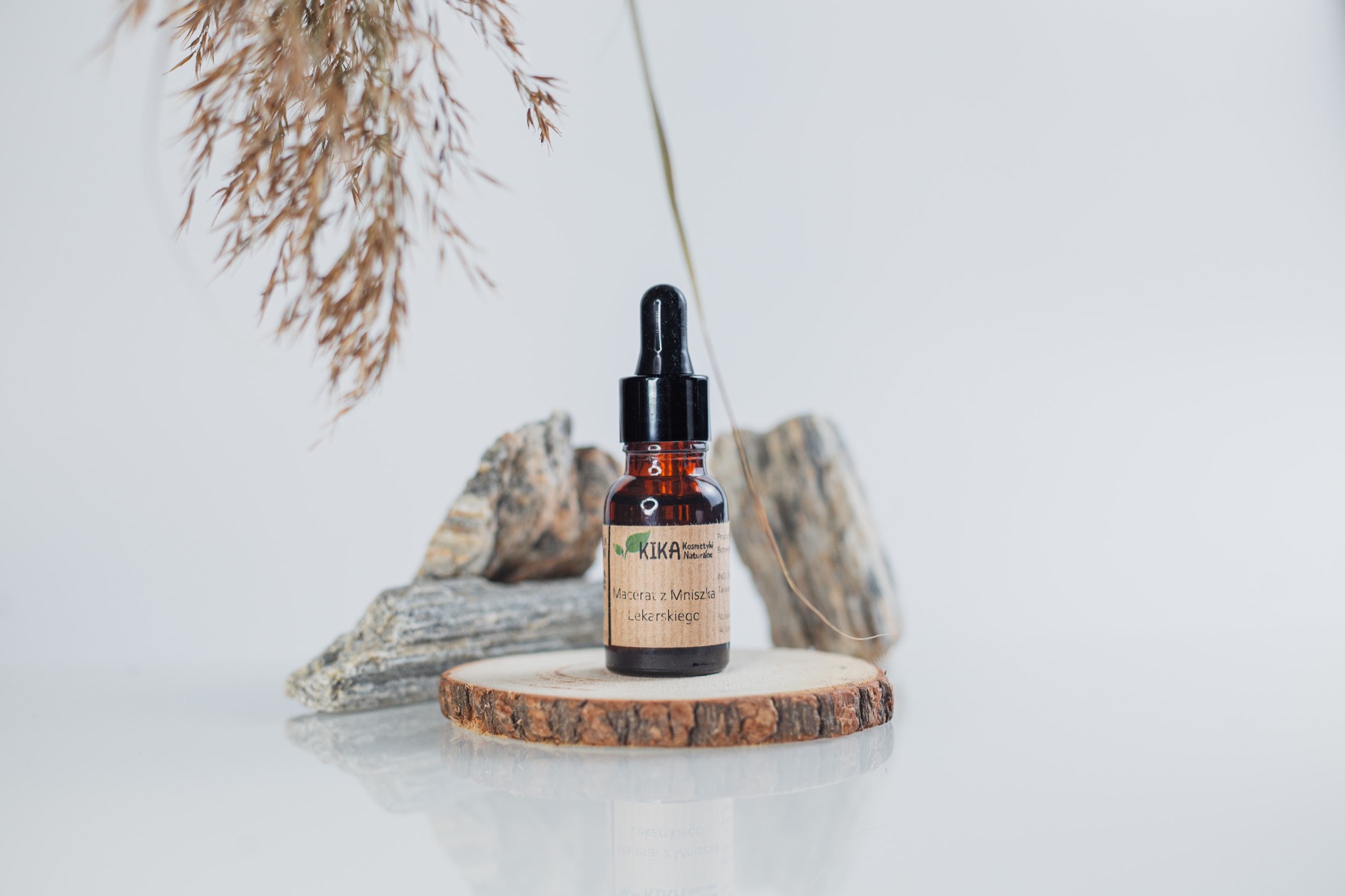 Brązowa buteleczka z pipetą, etykietą 'KIKA Kosmetyki Naturalne', umieszczona na drewnianym krążku, z kamieniami w tle i gałązką trawy, prezentacja produktu na białym tle.