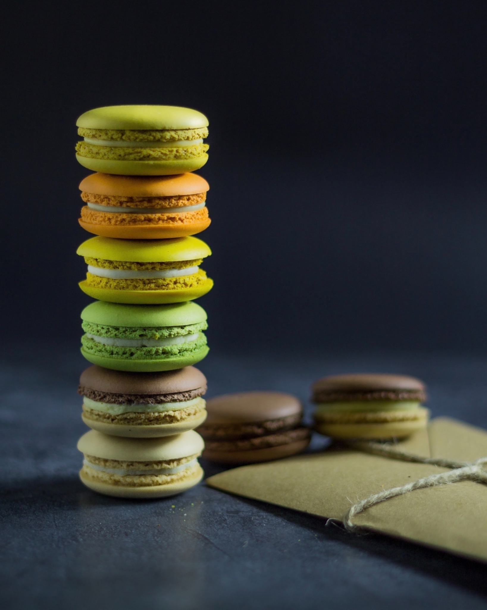 Wieża z kolorowych makaroników na ciemnym tle, obok leżą dwa makaroniki i brązowa koperta związana sznurkiem.