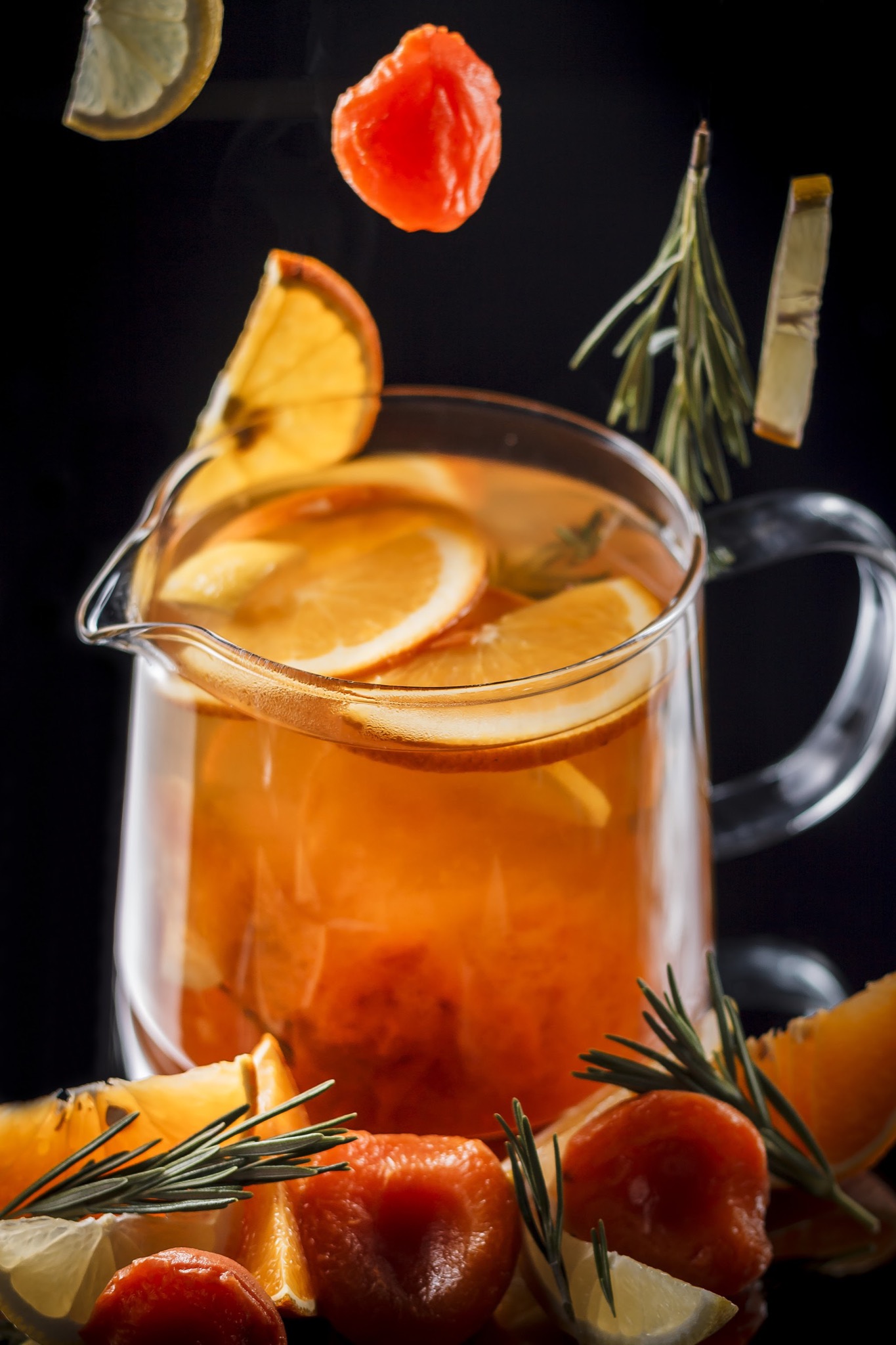 Szklany dzbanek z napojem pomarańczowym, plastrami pomarańczy i cytryny, gałązkami rozmarynu, unoszącymi się kawałkami owoców na czarnym tle.