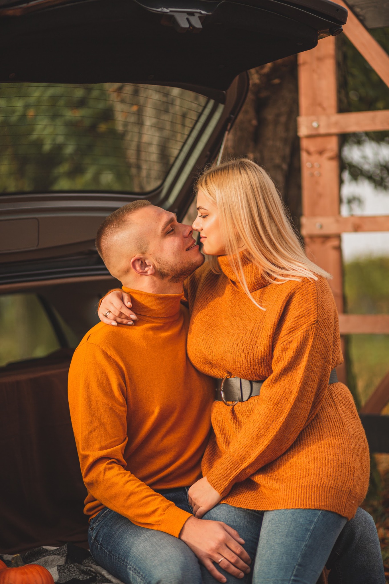 Zakochana para w pomarańczowych swetrach siedzi w bagażniku samochodu na tle jesiennego krajobrazu, bliskość twarzy, zaręczynowy pierścionek na palcu kobiety.