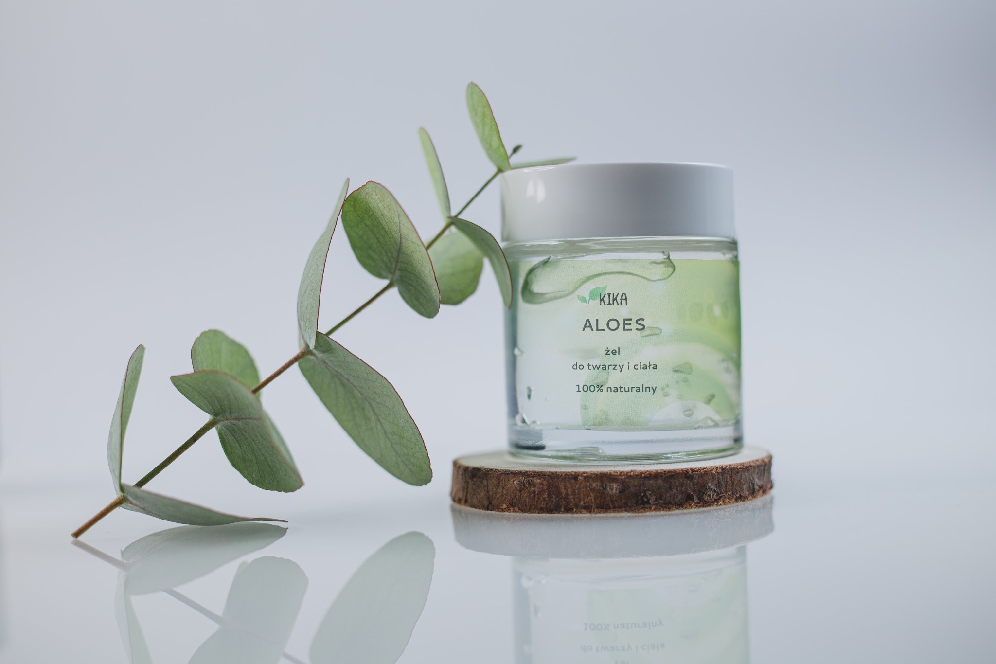 Szklany słoiczek z żelem aloesowym marki Kika Aloes na drewnianym podkładce, obok gałązka eukaliptusa, całość na białym tle z odbiciem.