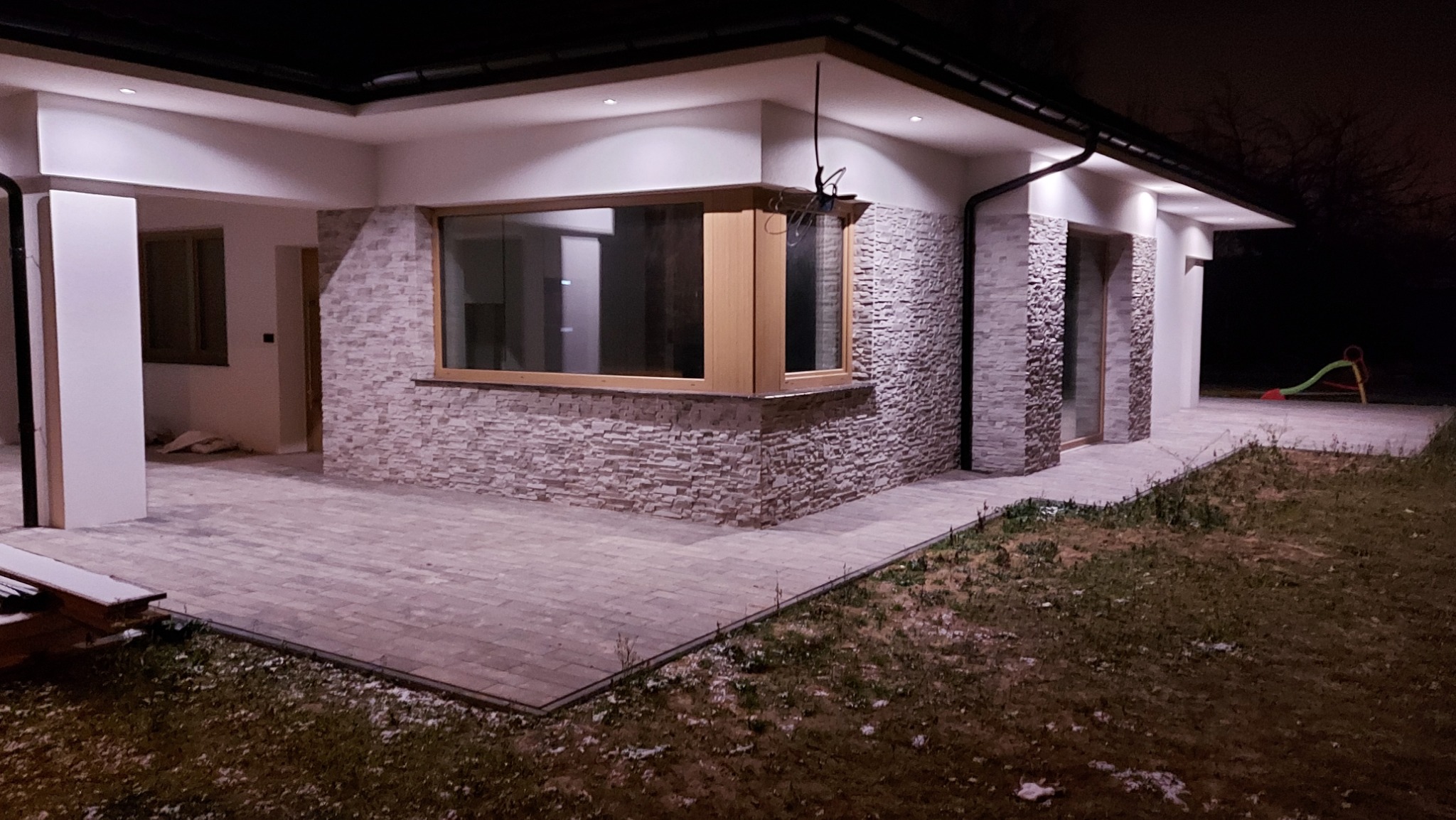 Wykończona elewacja domu jednorodzinnego w Lublinie z dekoracyjnym kamieniem elewacyjnym i oknami z drewnianą stolarką, oświetlona zewnętrznymi lampami.