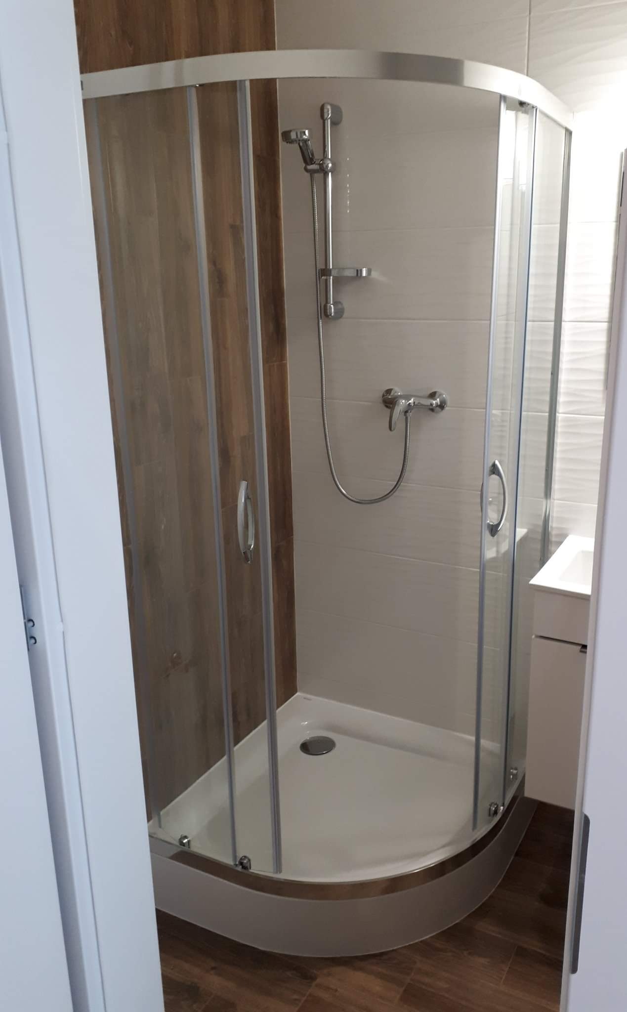 Nowoczesna kabina prysznicowa narożna z drewnopodobną ścianą i białymi płytkami, widok zza uchylonych drzwi łazienki.