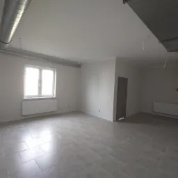 Pusty, jasny pokój z szarymi płytkami podłogowymi, białymi ścianami, grzejnikiem pod oknem, metalowymi rurami wentylacyjnymi pod sufitem i drzwiami w rogu