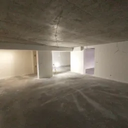 Surowe, betonowe wnętrze w trakcie prac wykończeniowych; ściany częściowo pokryte jasną warstwą, widoczne przewody elektryczne zwisające z sufitu, oświetlenie robocze na statywie w głębi...