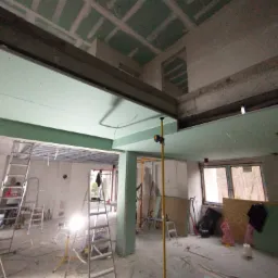 Wnętrze w trakcie montażu płyt kartonowo-gipsowych na suficie i ścianach, widoczne profile metalowe, drabiny i lampa budowlana na statywie.