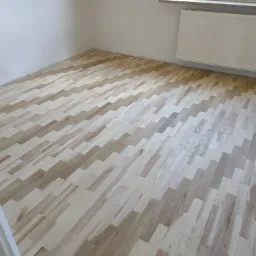 Nowo ułożona, jasna podłoga z parkietu w pustym pokoju, widoczne ściany pomalowane na biało i grzejnik.