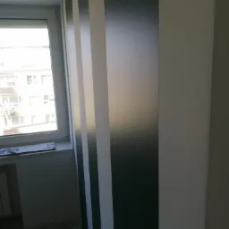 Pomalowana ściana w pionowe pasy: ciemnozielony, biały i jasnoszary, widok z boku okna z parapetem, na którym leży kartonowe opakowanie.