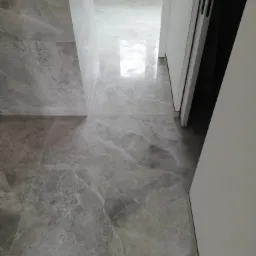 Wykończona podłoga w łazience z szarych płytek imitujących marmur, z widoczną białą ścianą i otwartymi drzwiami przesuwnymi.