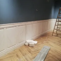 Pokój w trakcie wykańczania: ściany pomalowane na ciemny niebieski kolor z zamontowaną białą boazerią imitującą marmur, drewniana podłoga ułożona w jodełkę, fragment drabiny drewnianej w rogu,...