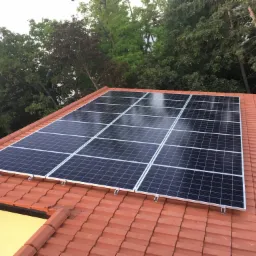 Panele słoneczne zamontowane na dachu z czerwonej dachówki, widok z góry, w tle drzewa.