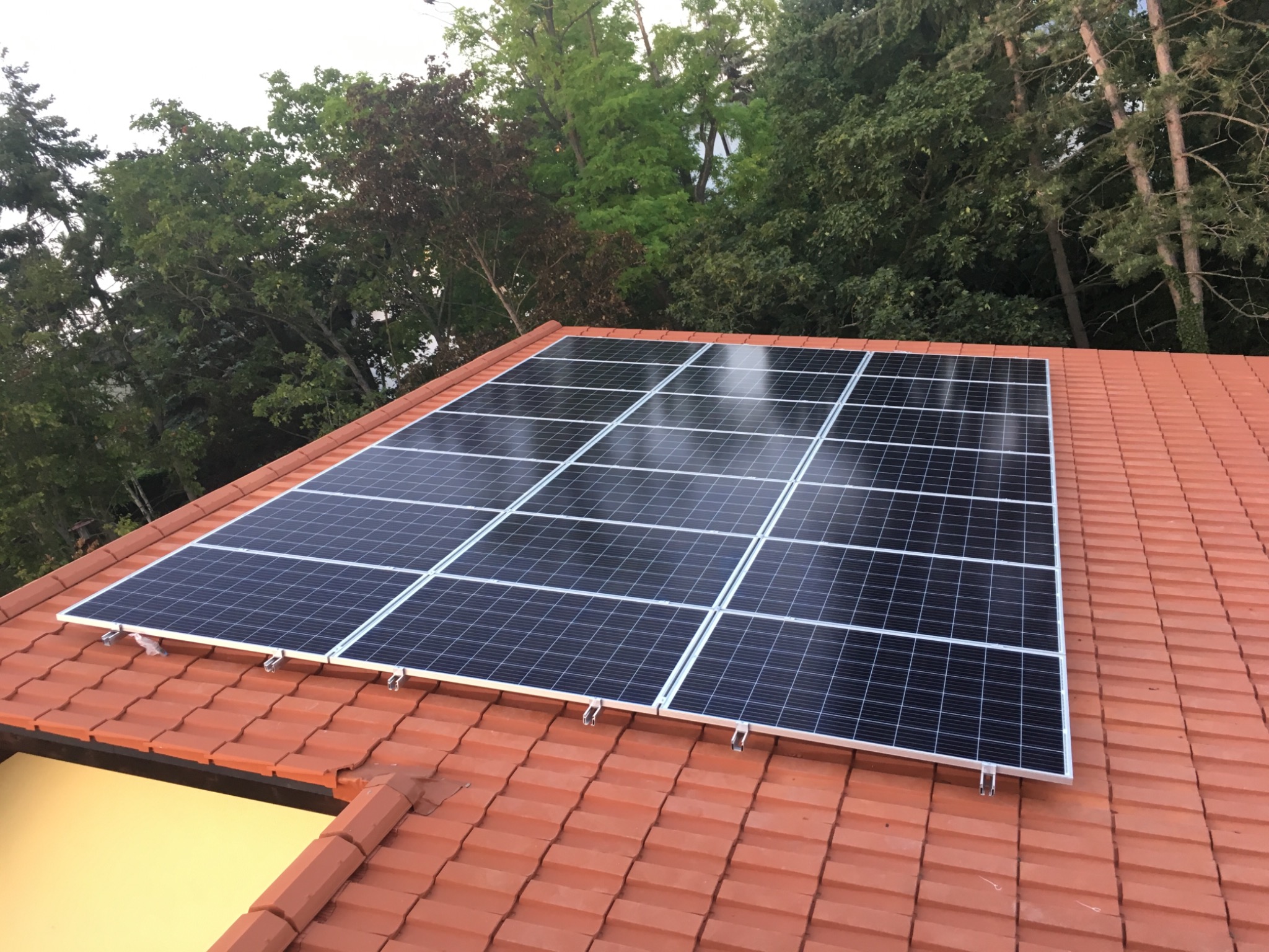 Panele słoneczne zamontowane na dachu z czerwonej dachówki, widok z góry, w tle drzewa.