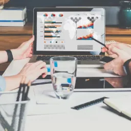 Dwie osoby analizują dane na laptopie, wskazując na wykresy słupkowe i mapę świata, szklanka wody, notatnik i ołówki na biurku.