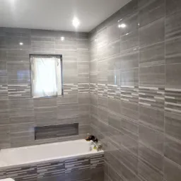 Wykończona łazienka z szarymi, poziomymi płytkami imitującymi kamień, wstawki z mozaiki, biała wanna, okno z zasłoniętą szybą i wnęka nad wanną.