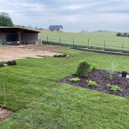 PRO GARDEN Marek Mazurek - Układanie trawy z rolki w ogrodzie, widoczne fragmenty ułożonej trawy, rolki trawy, przygotowany teren i rabata z nasadzeniami, w tle budowla ogrodowa i krajobraz wiejski.