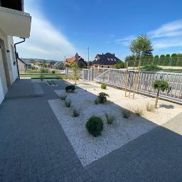 PRO GARDEN Marek Mazurek - Nowoczesny ogród przydomowy z jasnym żwirem, niskimi krzewami ozdobnymi, trawami i brukowaną ścieżką prowadzącą do domu z widokiem na okoliczne domy w tle.