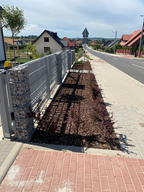 Nowoczesne ogrodzenie gabionowe z metalowymi przęsłami i nasadzeniami z kory i purpurowych krzewów wzdłuż chodnika przy ulicy w słoneczny dzień.