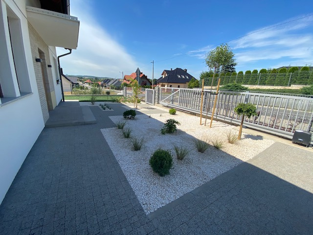 Nowoczesny ogród przydomowy z jasnym żwirem, niskimi krzewami ozdobnymi, trawami i brukowaną ścieżką prowadzącą do domu z widokiem na okoliczne domy w tle.