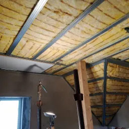 Montaż izolacji z wełny mineralnej w konstrukcji metalowej poddasza, widoczne fragmenty płyty kartonowo-gipsowej, narzędzia i lampa budowlana.