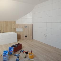 FIRMA DOCIEPLENIOWO WYKOŃCZENIOWA TOMASZ GURSKI - Wykończenie łazienki na poddaszu: białe płytki ścienne z poziomym wzorem, zabudowa wanny i stelaż WC w okleinie drewnopodobnej, worki z klejem, wiadro i narzędzia na podłodze, tymczasowe oświetlenie.