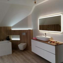 FIRMA DOCIEPLENIOWO WYKOŃCZENIOWA TOMASZ GURSKI - Nowoczesna łazienka na poddaszu z oświetlonym lustrem, białą umywalką nablatową, wiszącą toaletą i drewnianymi panelami ściennymi, w trakcie wykańczania, widoczne narzędzia na blacie.
