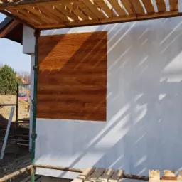 Fragment elewacji budynku z dekoracyjnym tynkiem imitującym drewno, widoczne rusztowanie i koparka w tle.