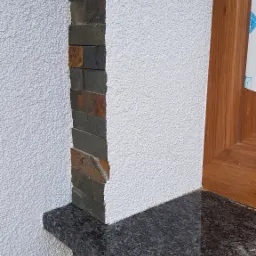 Fragment elewacji z białym tynkiem barankiem, obłożony dekoracyjnym kamieniem łupkowym w odcieniach szarości i brązu oraz ciemnoszarym parapetem z granitu przy drewnianej ramie okiennej.