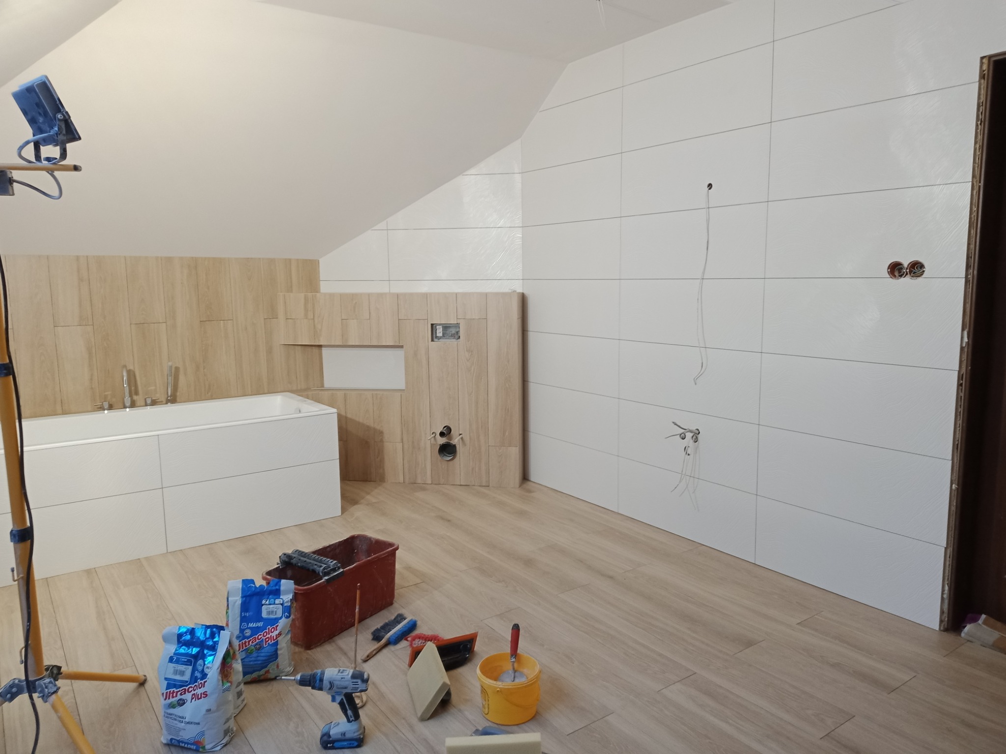 Wykończenie łazienki na poddaszu: białe płytki ścienne z poziomym wzorem, zabudowa wanny i stelaż WC w okleinie drewnopodobnej, worki z klejem, wiadro i narzędzia na podłodze, tymczasowe oświetlenie.