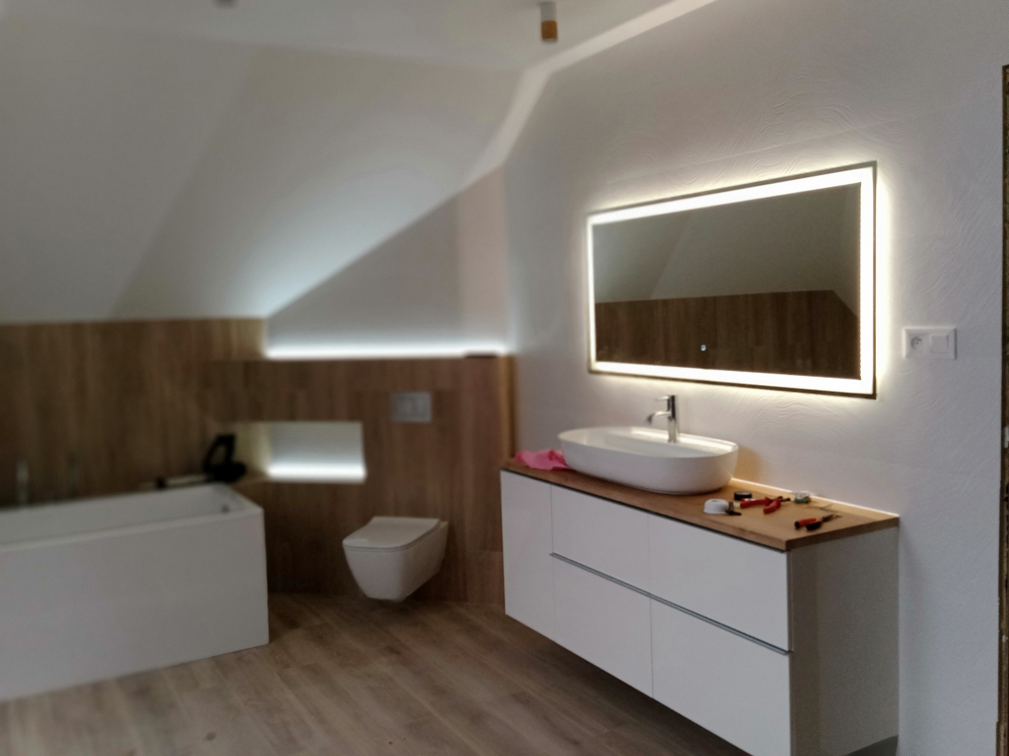 Nowoczesna łazienka na poddaszu z oświetlonym lustrem, białą umywalką nablatową, wiszącą toaletą i drewnianymi panelami ściennymi, w trakcie wykańczania, widoczne narzędzia na blacie.