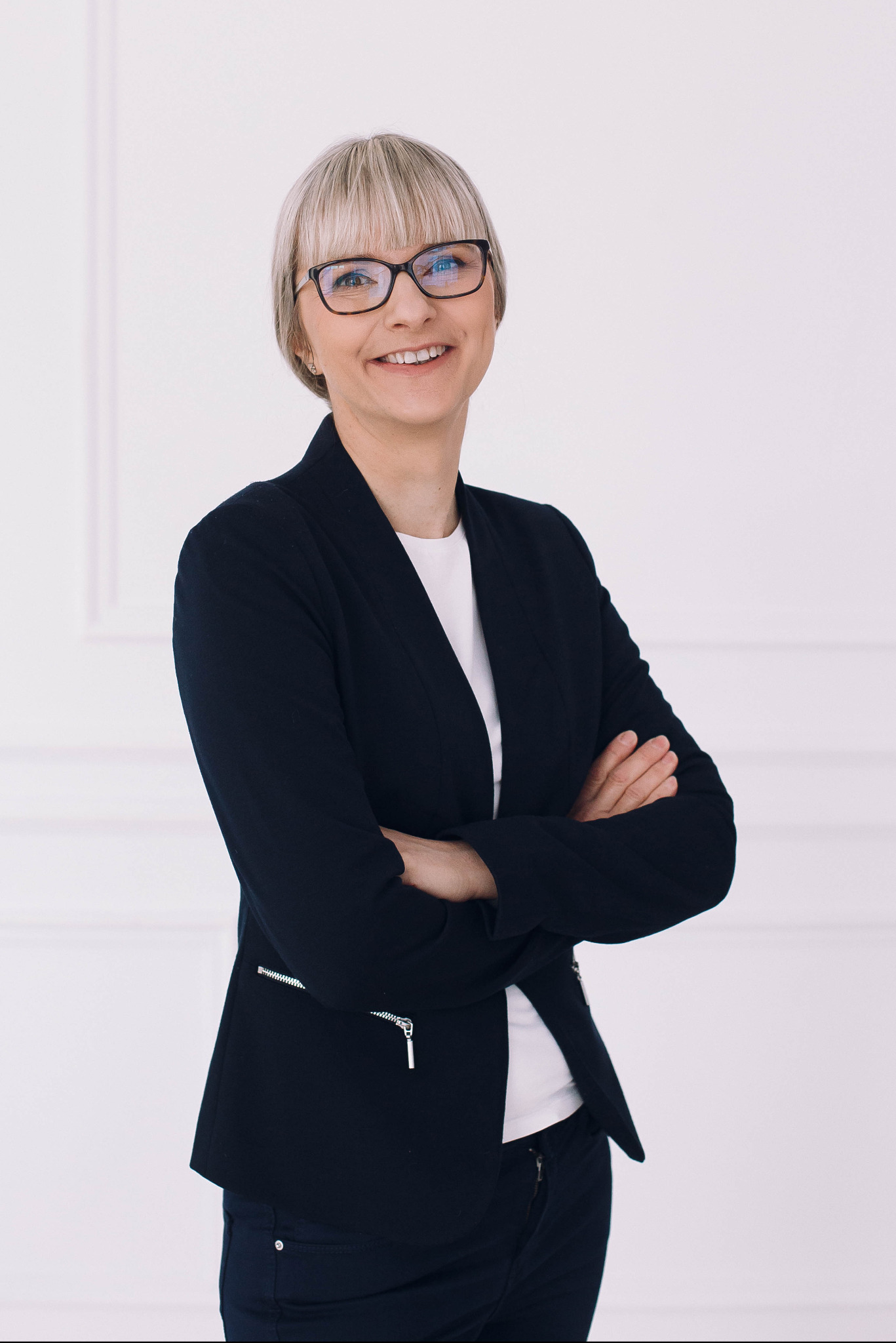 Uśmiechnięta kobieta w okularach i czarnej marynarce, z rękami skrzyżowanymi na piersiach, na tle białej ściany.