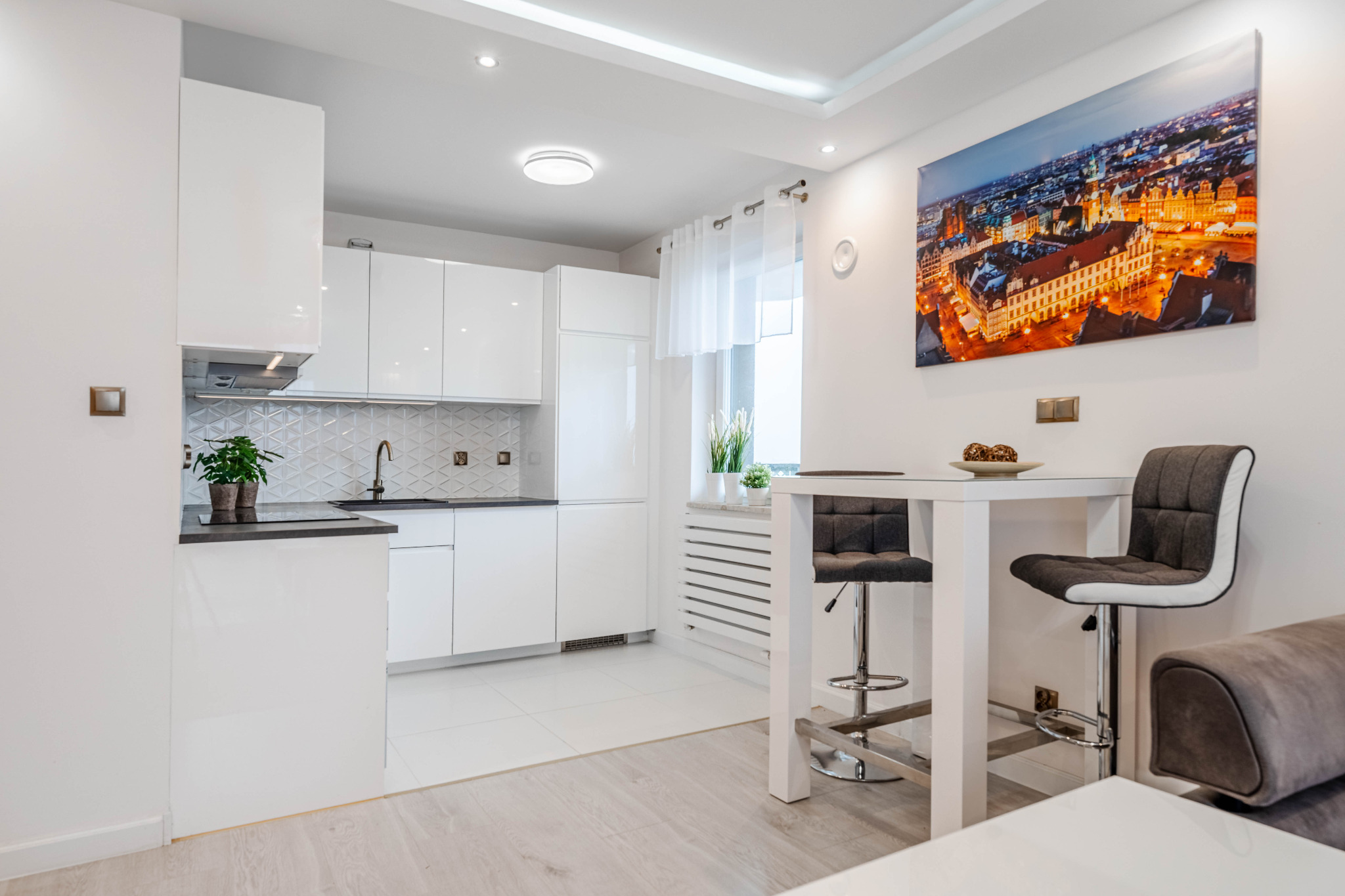 Jasna, nowoczesna kuchnia z białymi szafkami i czarnym blatem, uzupełniona drewnianą podłogą i obrazem panoramy miasta na ścianie.