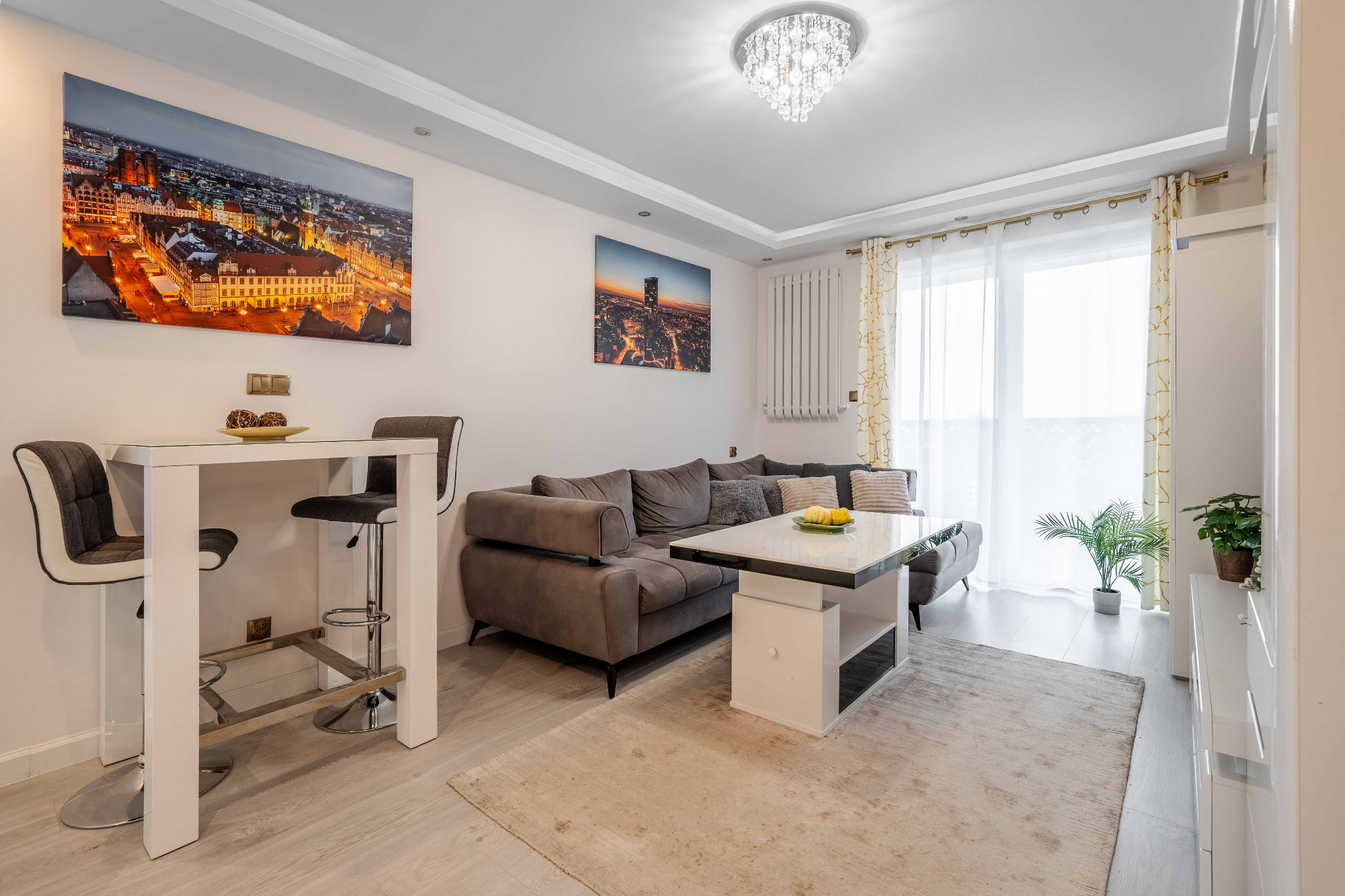 Jasne wnętrze salonu z szarą sofą, białym stolikiem kawowym i stołem barowym, dekorowane obrazami miast i kryształowym żyrandolem.