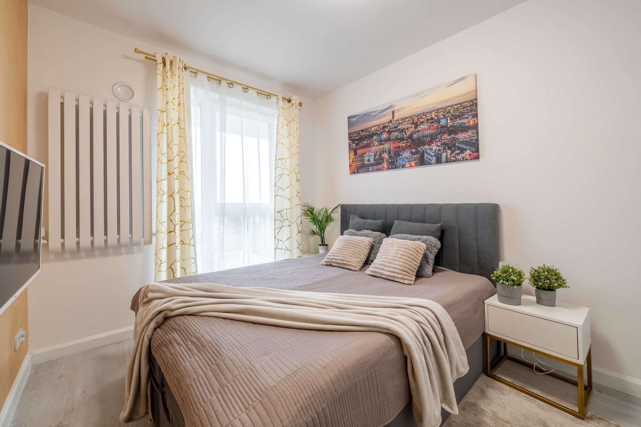 Jasna sypialnia z szarym łóżkiem, beżową narzutą i poduszkami, obraz z panoramą miasta nad łóżkiem, biała szafka nocna z roślinami w doniczkach, okno z białymi zasłonami i wzorzystymi zasłonami.
