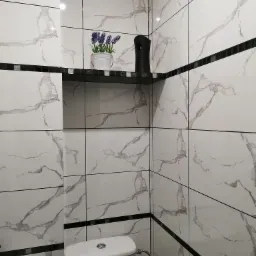 Wykończona łazienka z białymi płytkami imitującymi marmur, czarnymi akcentami w postaci listew i półki, na której stoi doniczka z lawendą i czarny odświeżacz powietrza. Widoczny fragment toalety...